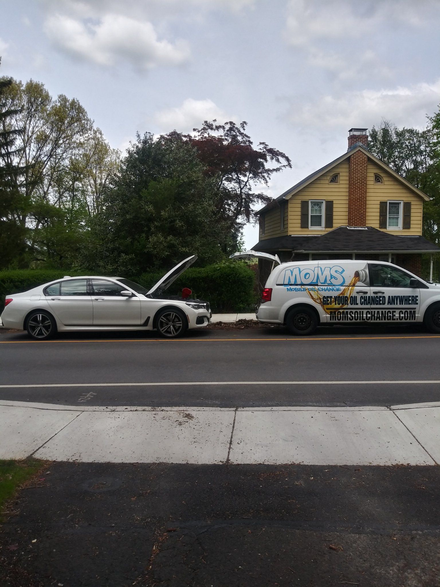 MOMS van beside a white BMW with its hood up during a full synthetic oil change in a customer's driveway.
